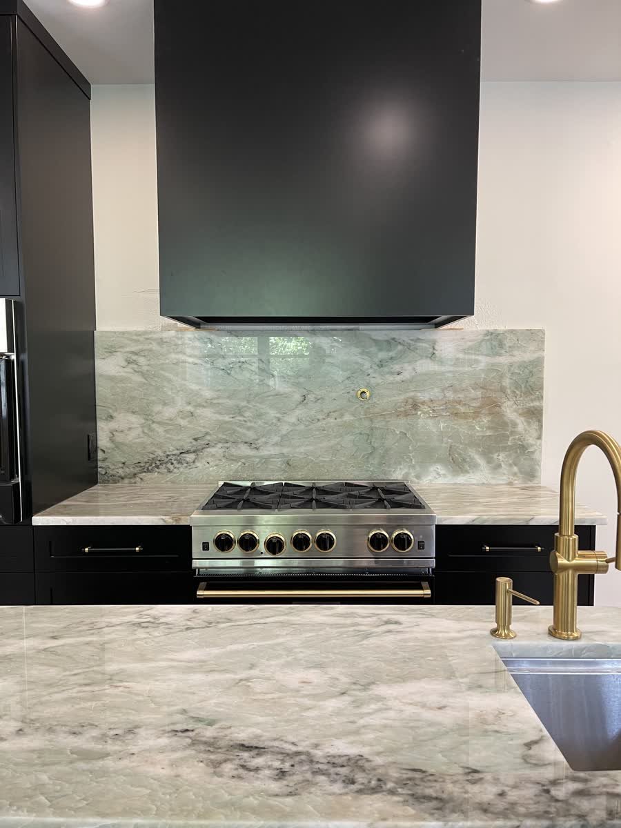 Black kitchen with a black range hood, a full-height stone backsplash and a brass faucet